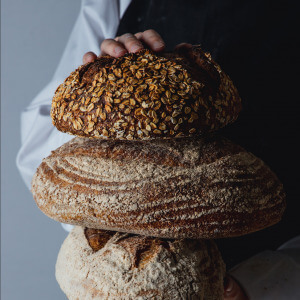 An image of a smartly dressed baker holding three different types of artisan bread.